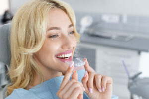 Happy young attractive blonde woman holding invisible braces or trainer while sitting at dental chair, closeup portrait