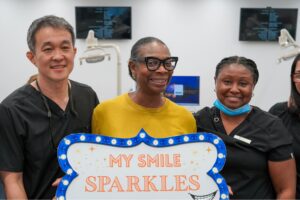 A smiling patient holding a "My Smile Sparkles" sign alongside a male dentist and a female assistant